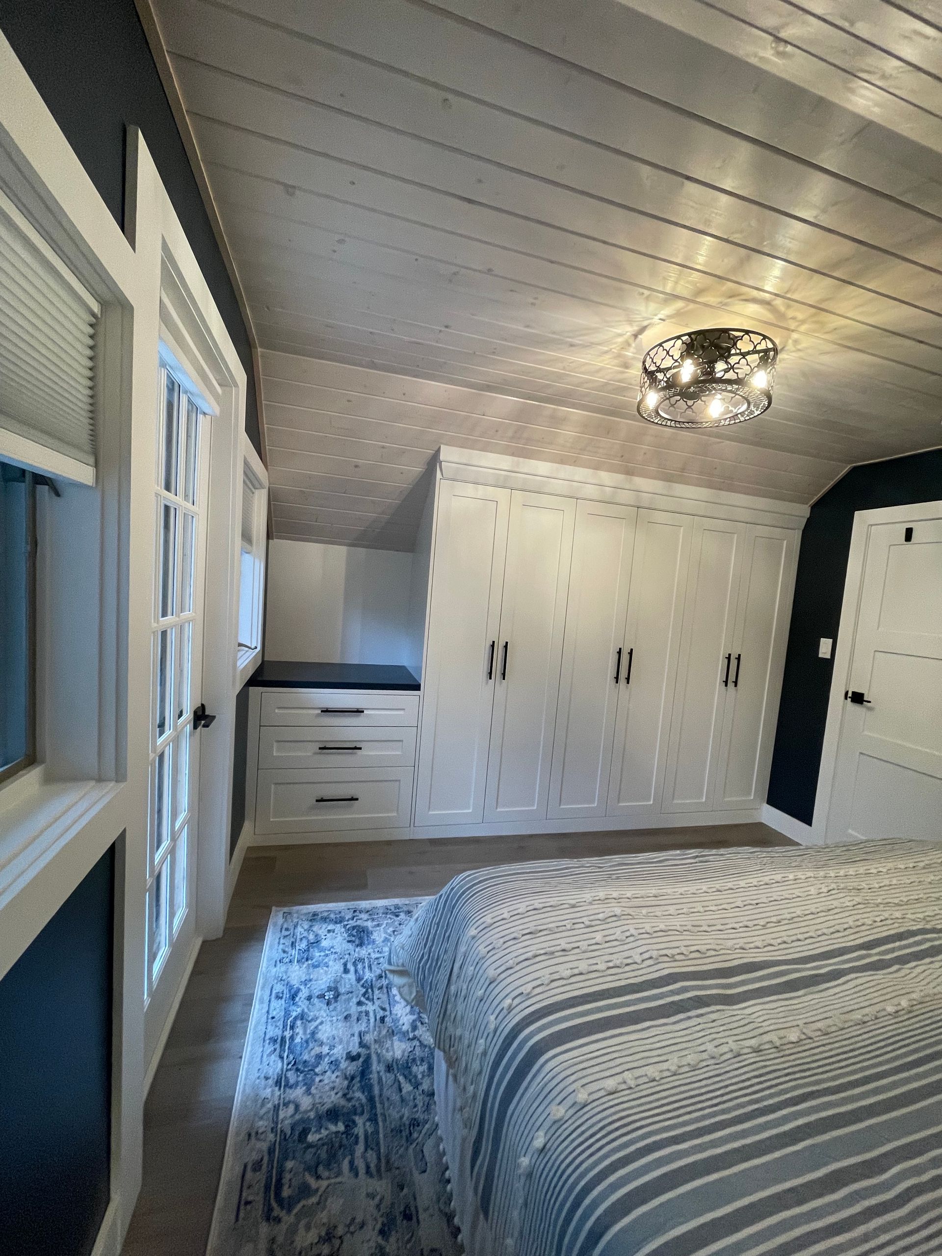 Bedroom with white built-in closets, window, and rug. Gray striped bedspread. Angled, wood-paneled ceiling.