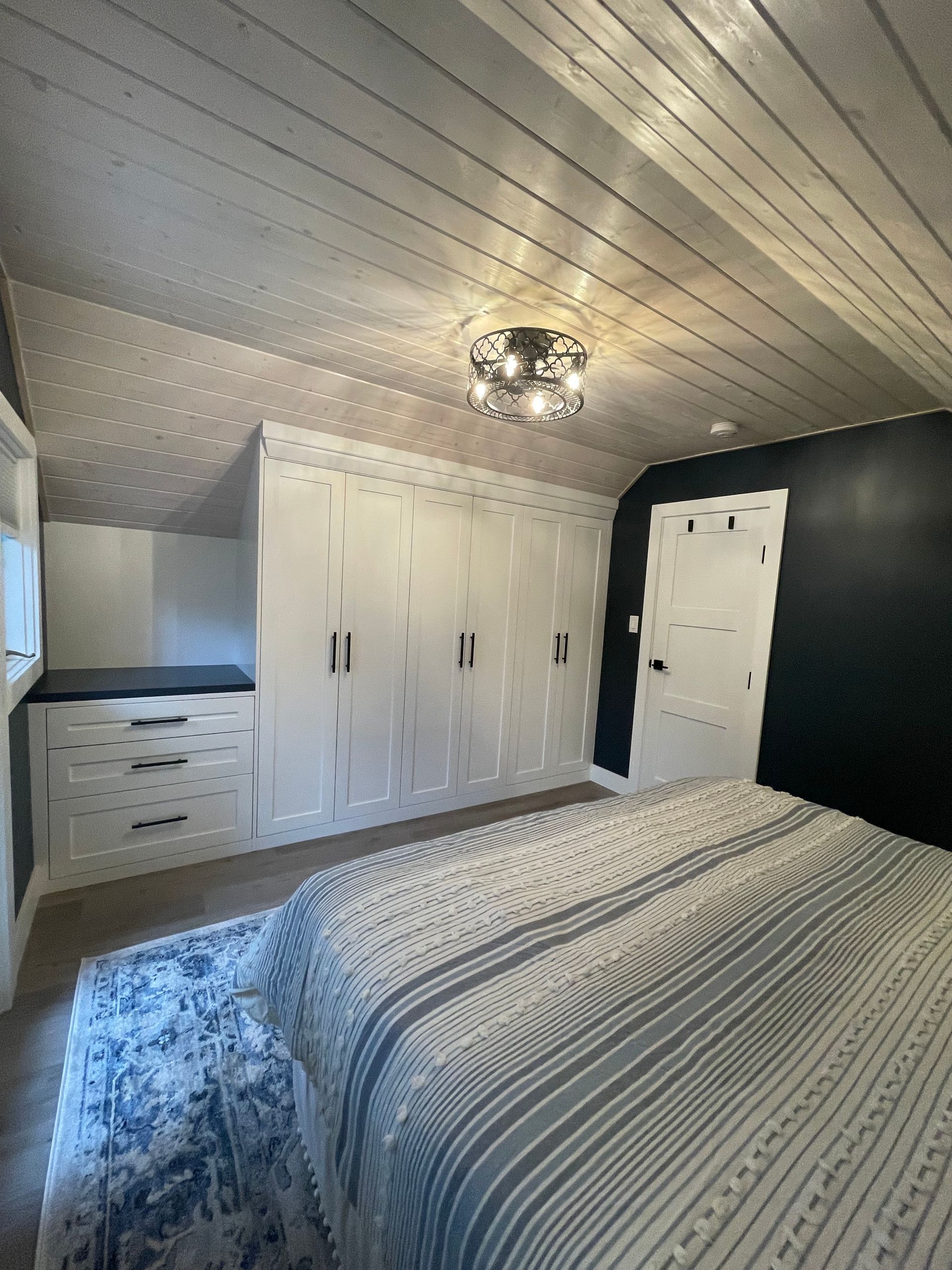 Bedroom with white built-in storage and black accent wall. Striped bedspread, wood ceiling, and area rug.