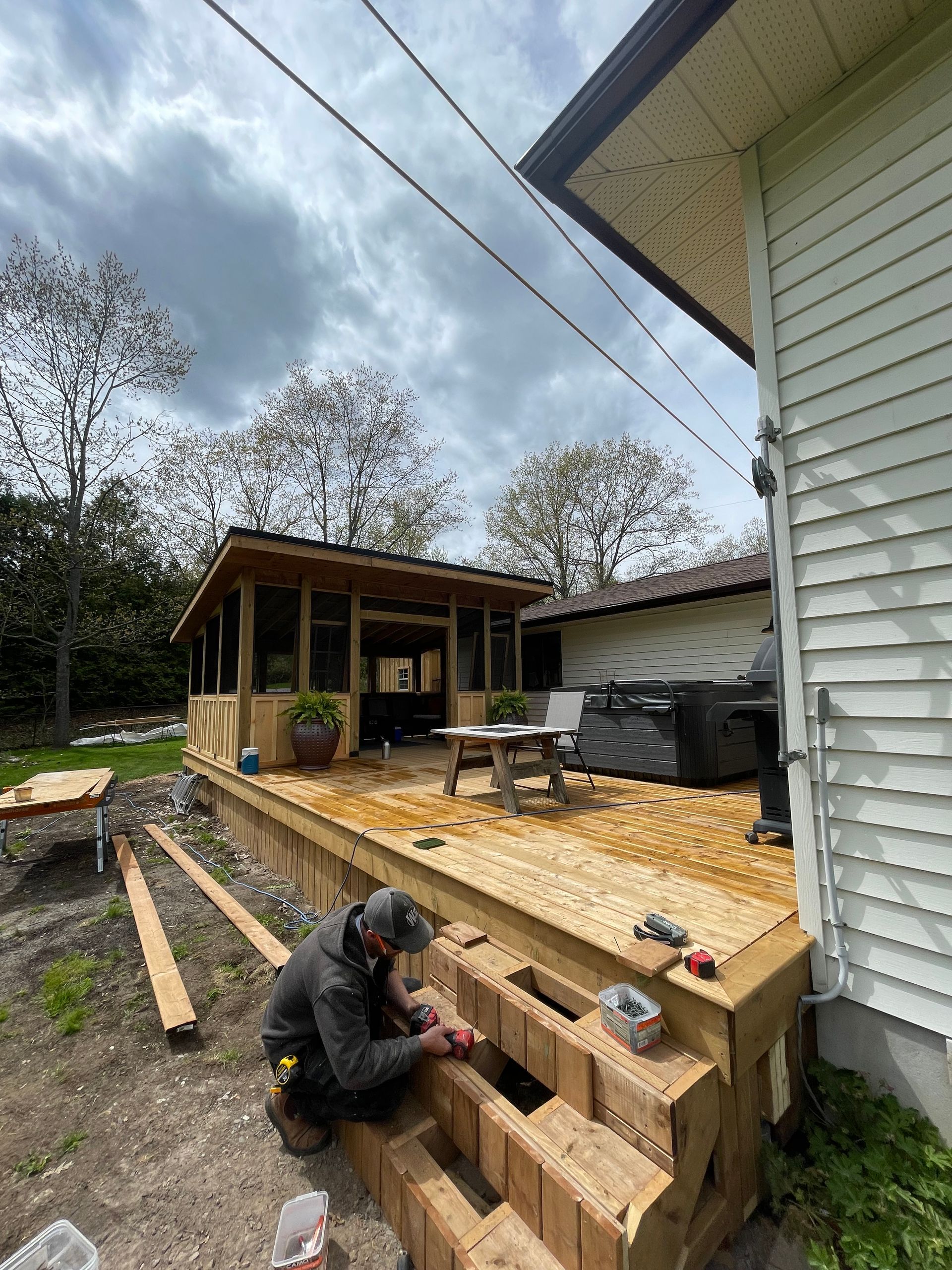 Man constructing wooden steps for a deck. A screened-in gazebo and house are in the background under a cloudy sky.