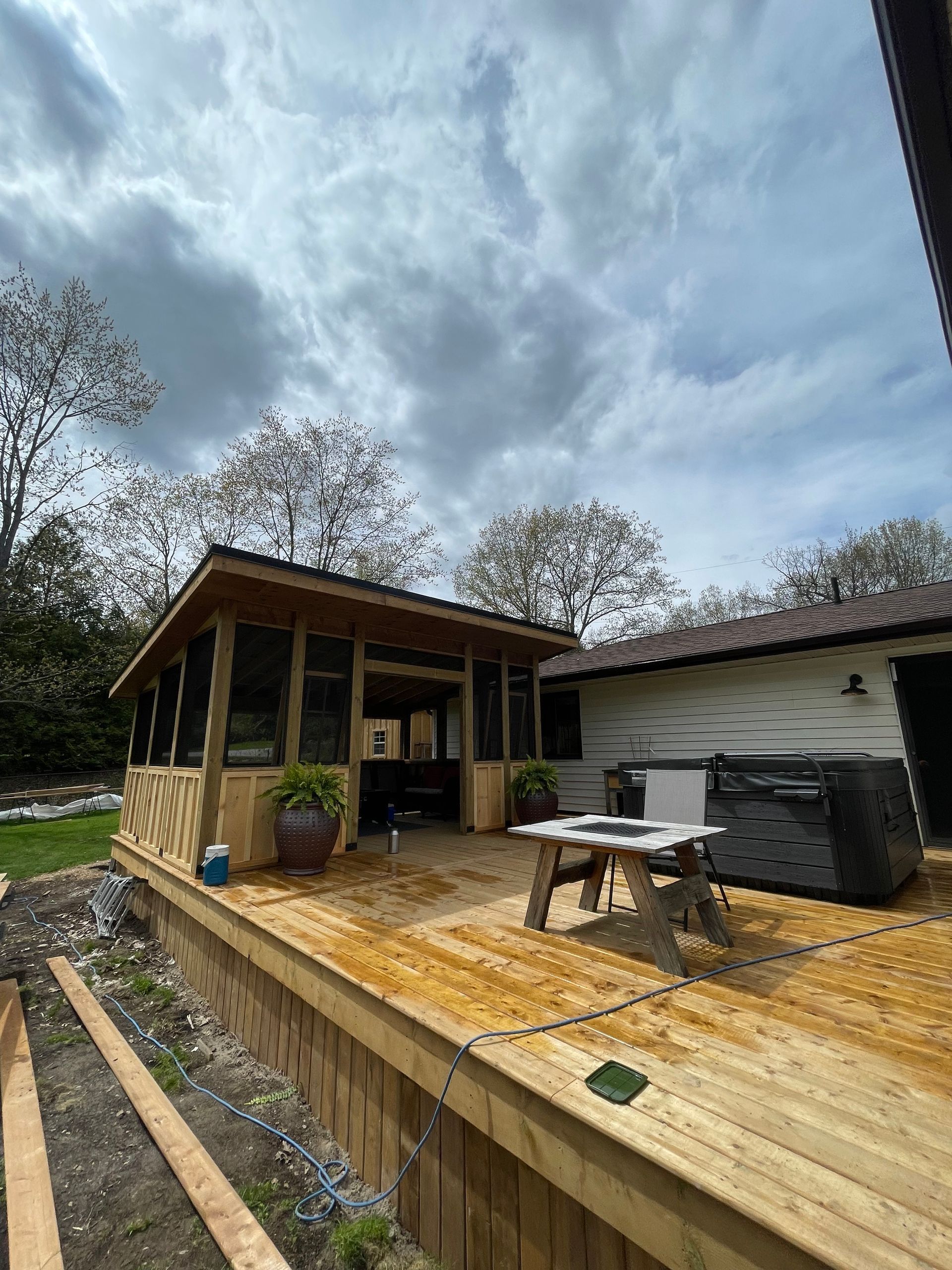 Wooden deck with screened-in gazebo, hot tub, and small table. Overcast sky in the background.