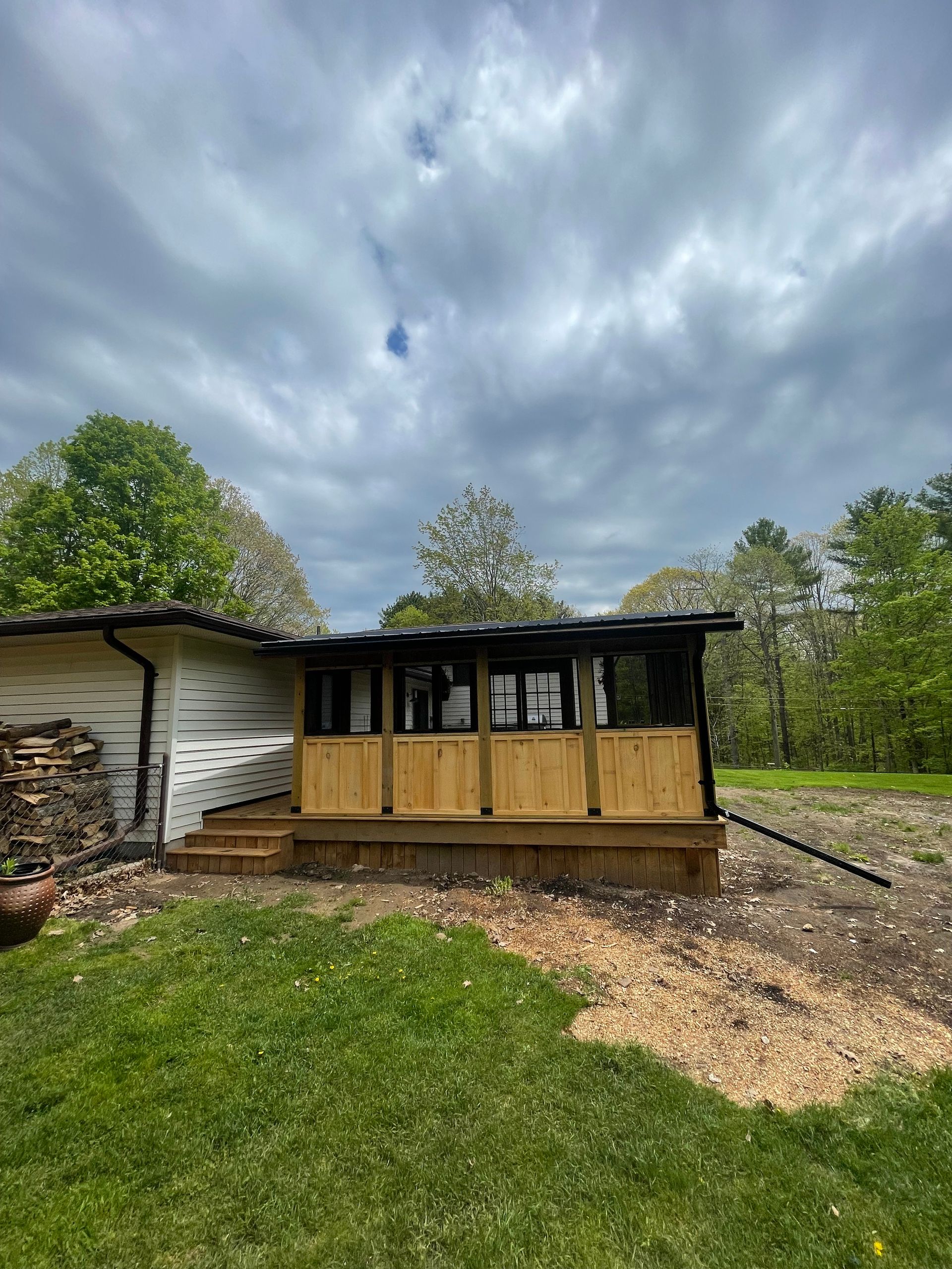 A wooden porch attached to a white building. The porch has a wooden railing and a screened enclosure, set against a cloudy sky.