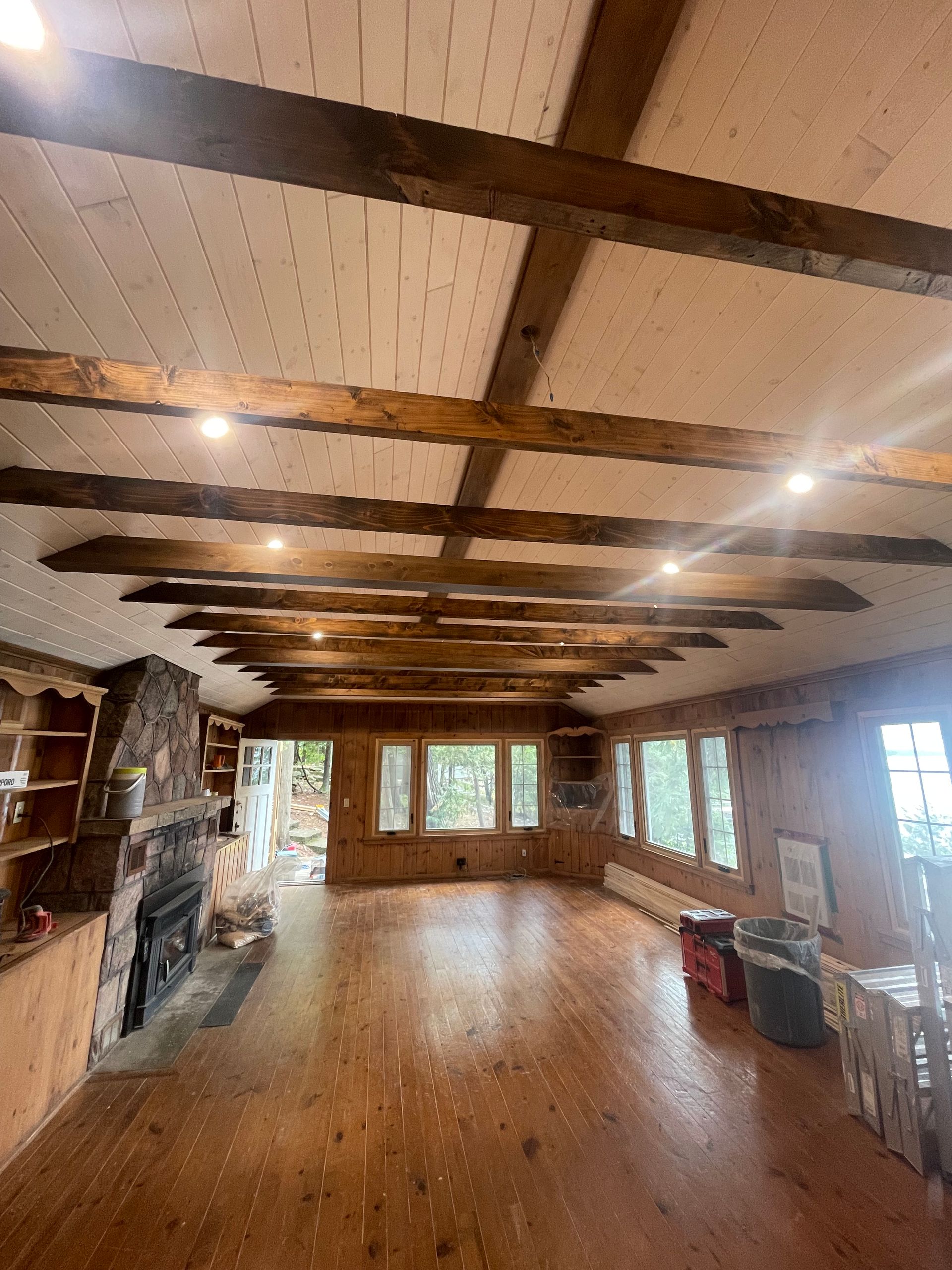 Rustic empty living room with wood floors, exposed beams, stone fireplace, and large windows.
