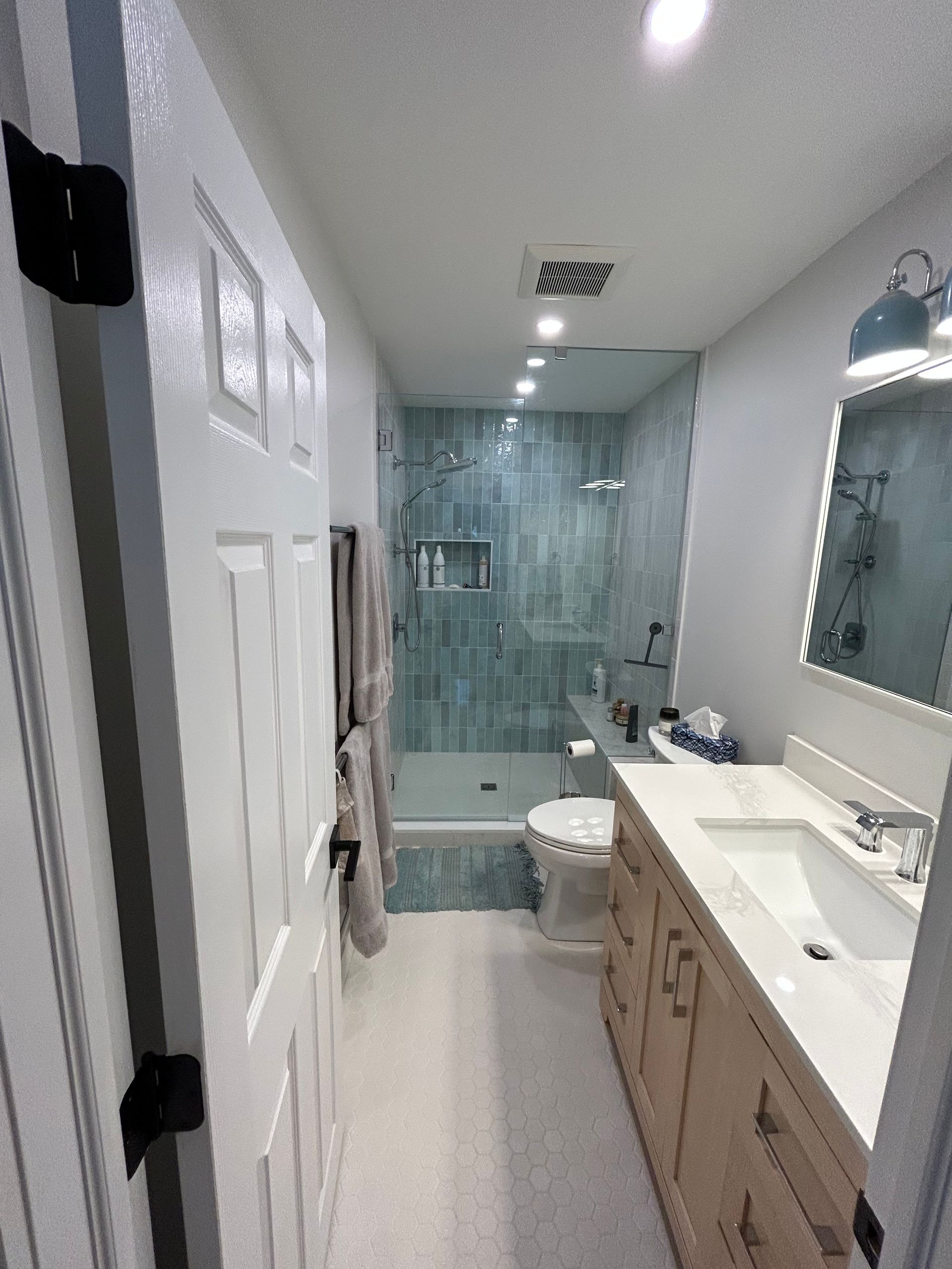 A long, narrow bathroom with a bathtub, toilet, and vanity. Teal tile in the shower area, white door.