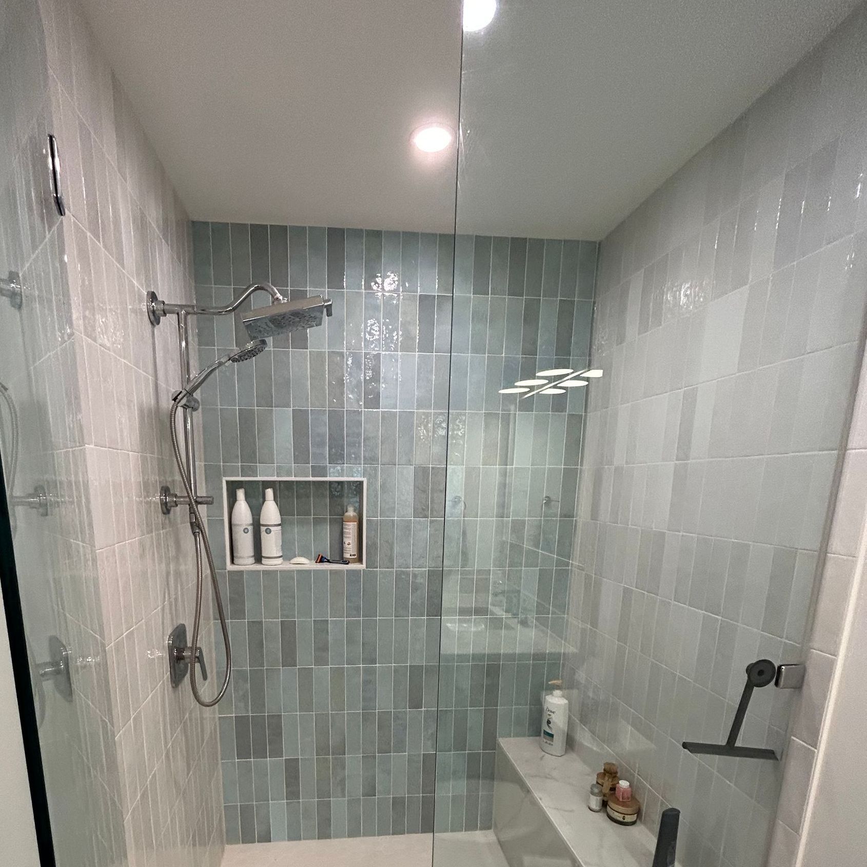 Bathroom shower with blue-green tiles, glass door, bench, and chrome fixtures.