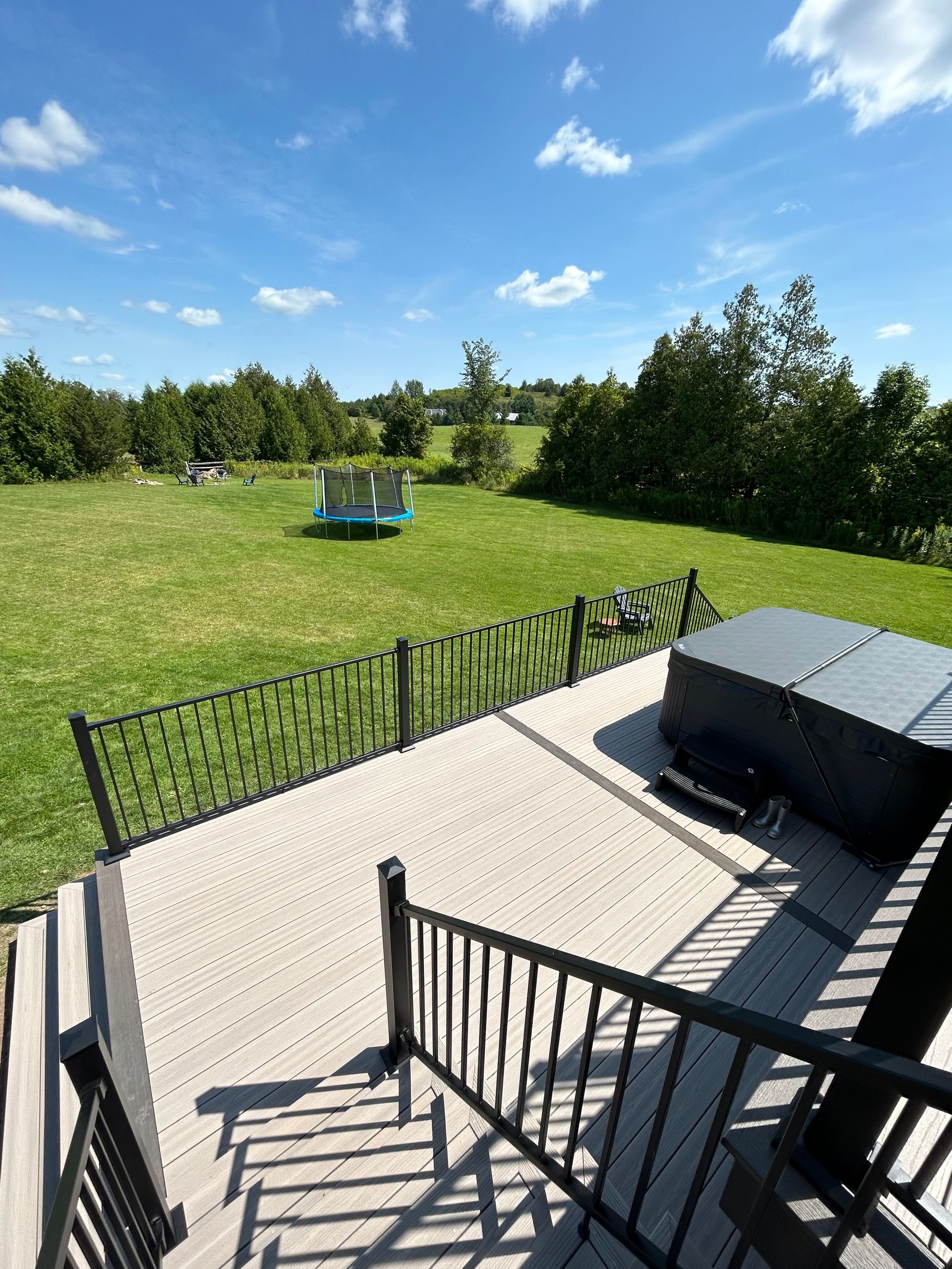 Wooden deck with a hot tub overlooking a grassy yard with trees and a trampoline under a blue sky.