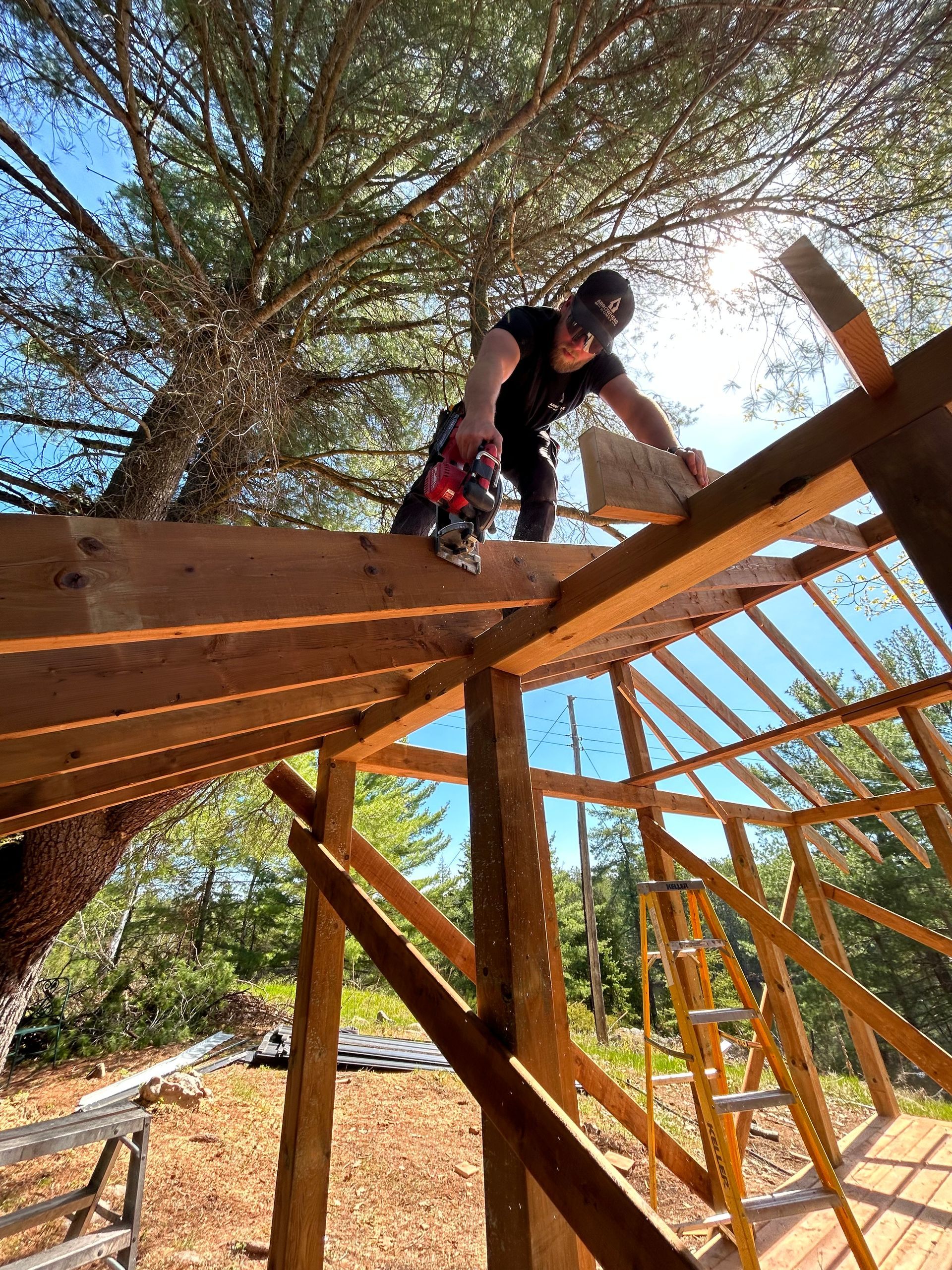 Person crouching on a wooden structure under trees, working on framing in bright sunlight