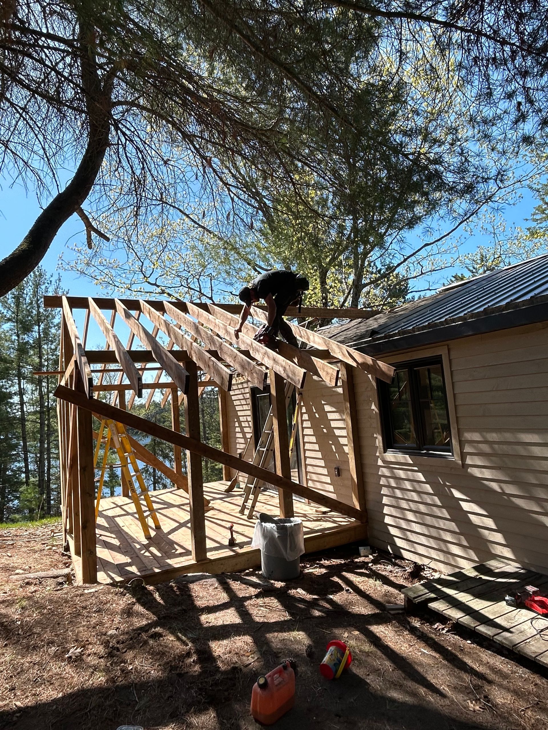 Person working on a wooden roof frame beside a house under tall trees