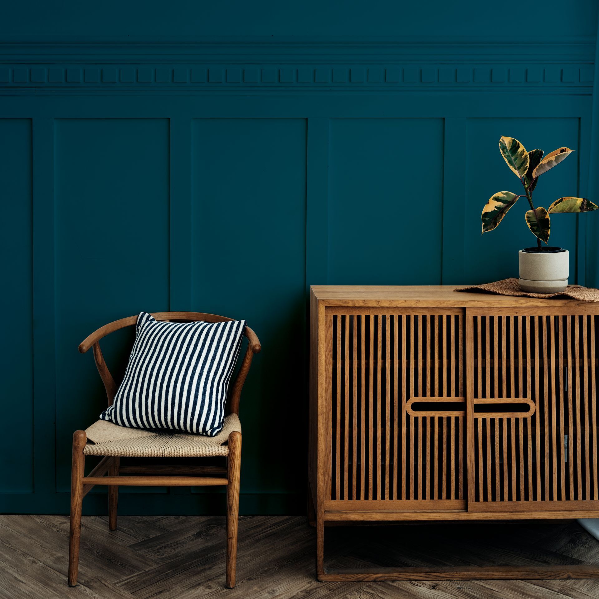 Teal paneled wall with a wooden chair, striped cushion, and cabinet with a potted plant.