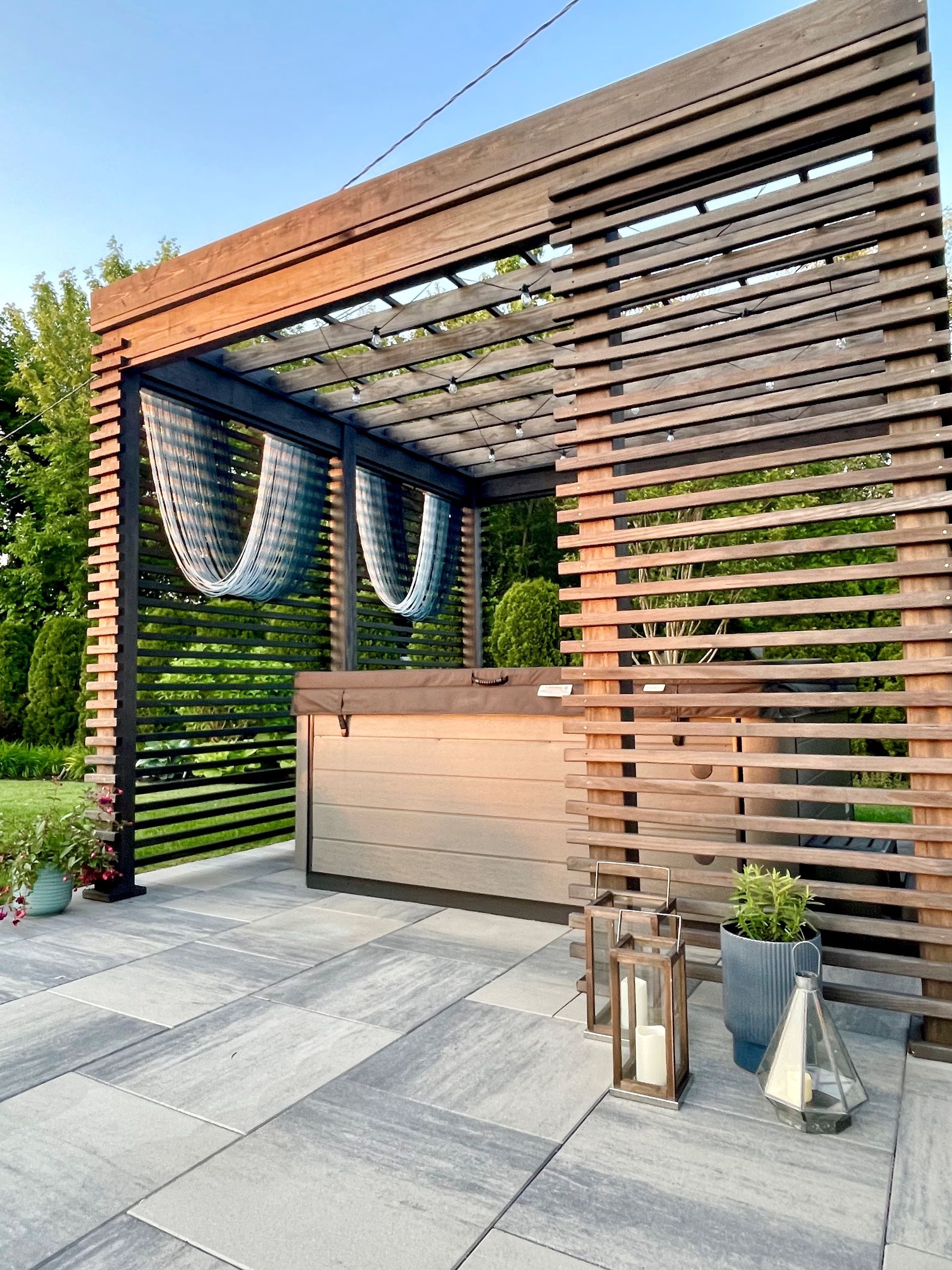 Wooden pergola with hot tub on a tiled patio, featuring a slatted privacy wall. Lanterns and plants add ambiance.