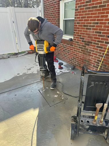 Worker using a jackhammer on a concrete patio beside a brick house