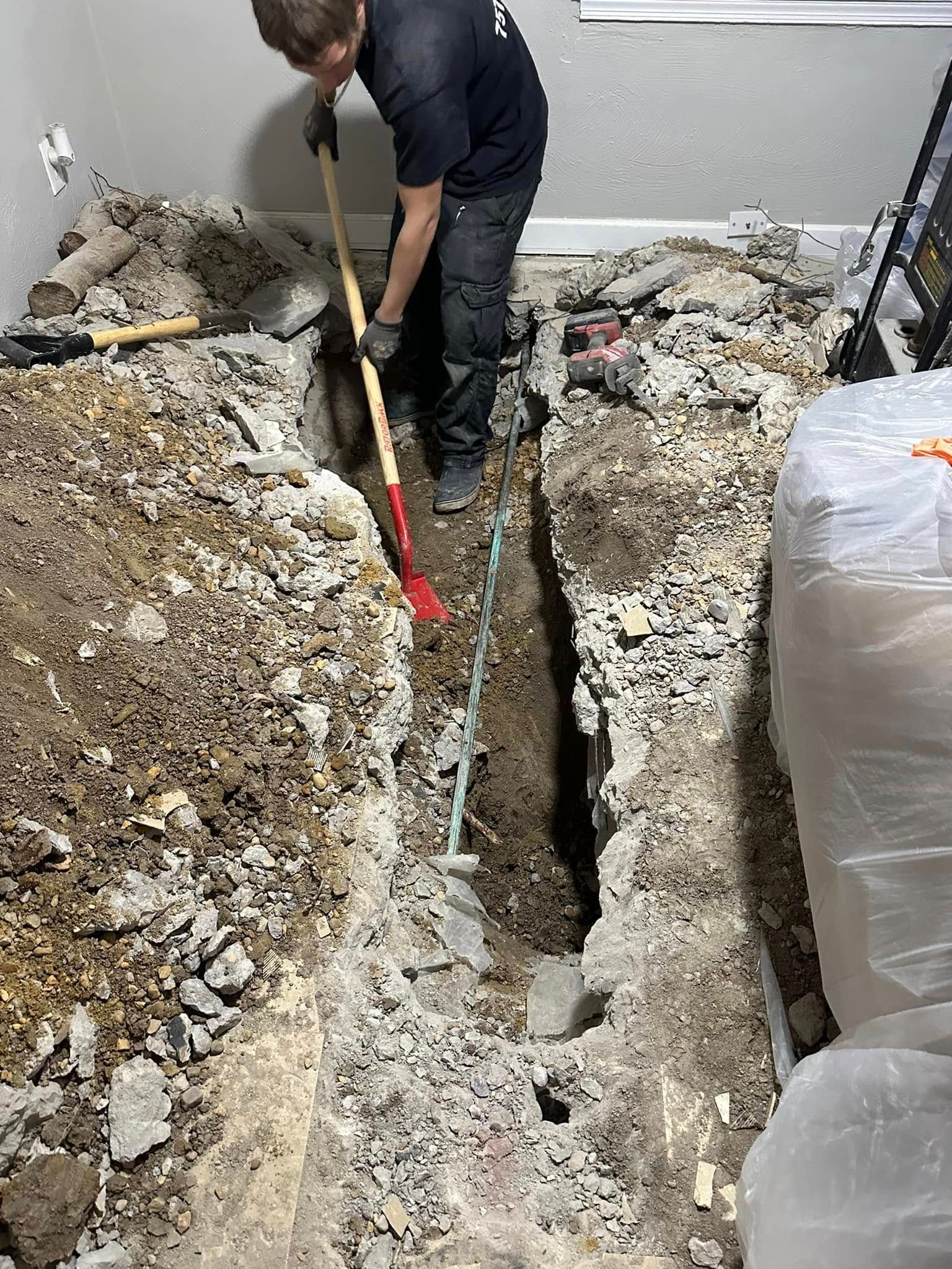 Worker digging a narrow trench in a debris-filled room with exposed dirt and insulation.