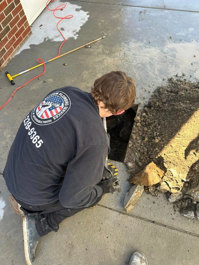 Worker kneeling by a sidewalk hole, clearing debris beside a chipped concrete curb and brick wall.