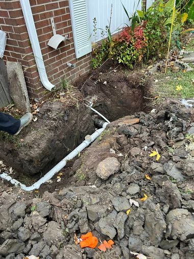 Dug trench beside a brick house with exposed white pipes and soil piled up near a garden bed.