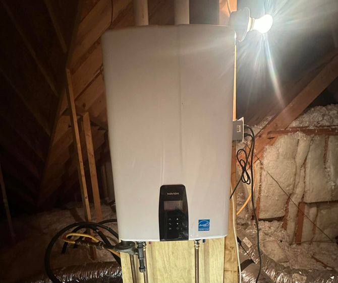 White wall-mounted furnace in an attic, with exposed insulation, wiring, and a bright work light nearby