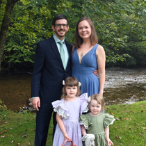 Family of four poses outdoors. Man in navy suit, woman in blue dress, two young girls in dresses stand on grass.