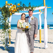 Couple smiles near bamboo wedding arch decorated with flowers on a beach with blue sky and ocean.