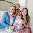 Family of four smiling, sitting on a bed. Mother holds baby, older daughter hugs father.