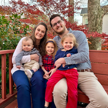Family of five smiling, sitting on a wooden bench outdoors. Red, blue, and brown clothing; fall foliage in background.
