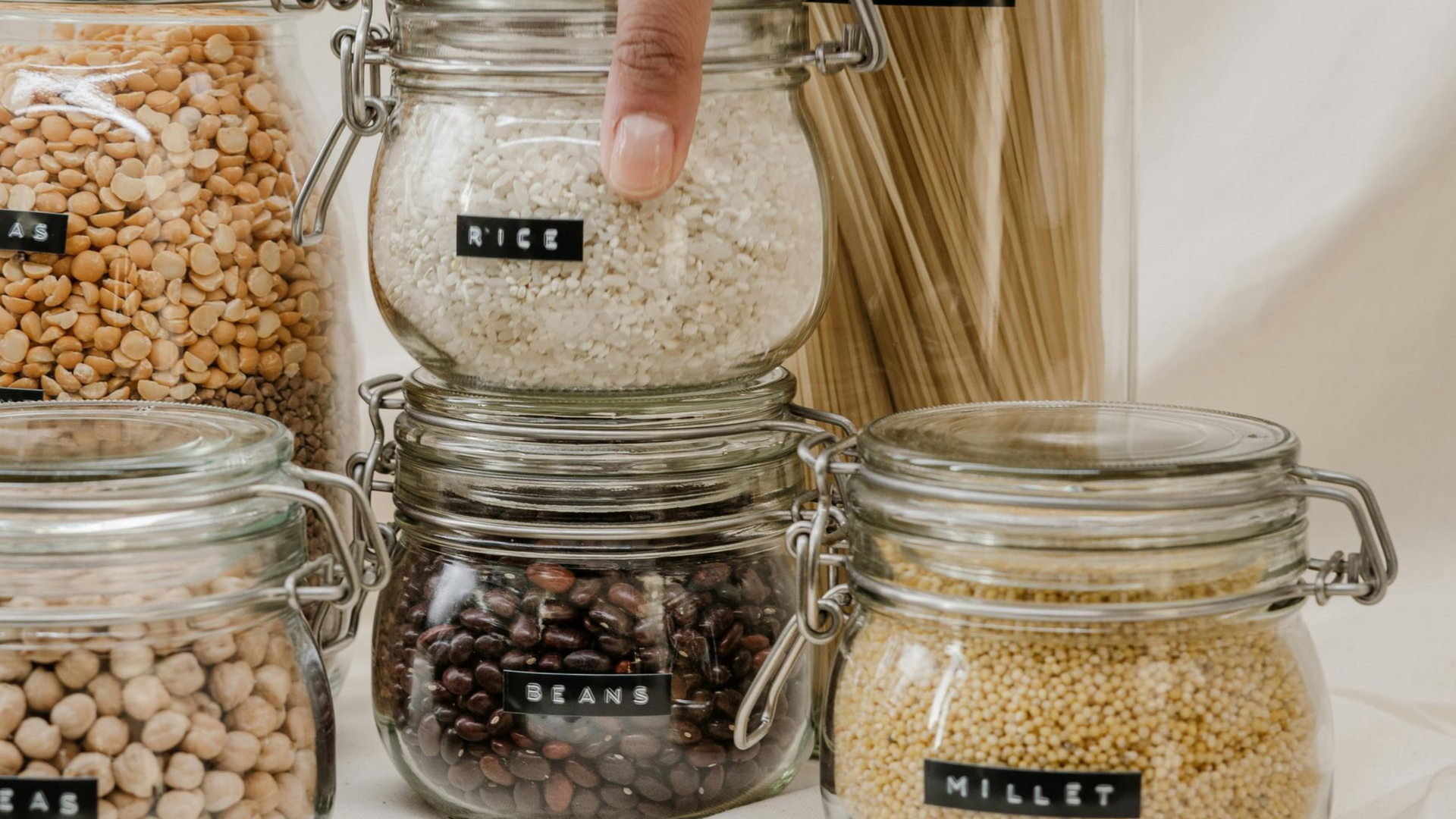 Person's finger pointing at a jar of rice. Jars filled with grains and beans neatly organized with labels.
