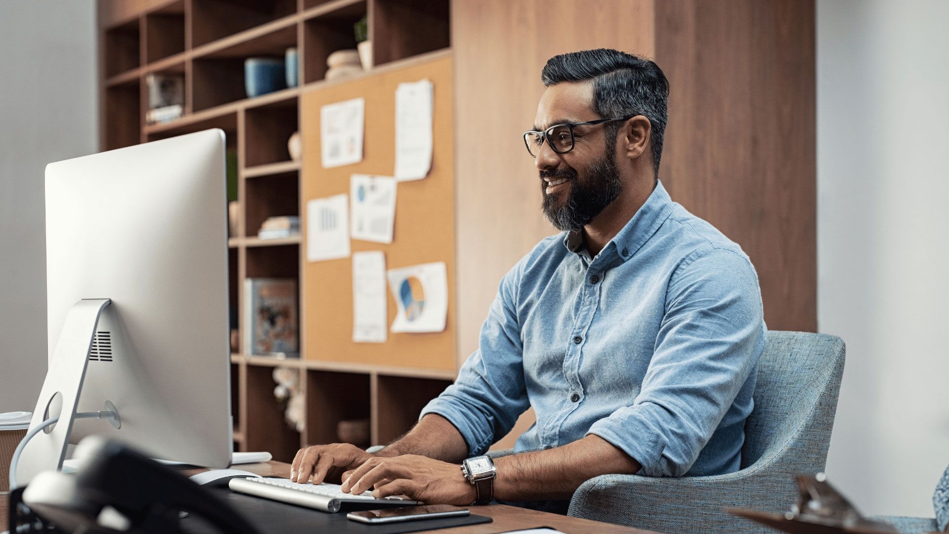 Man typing at a desk in a modern office, using a large desktop monitor and smiling slightly.