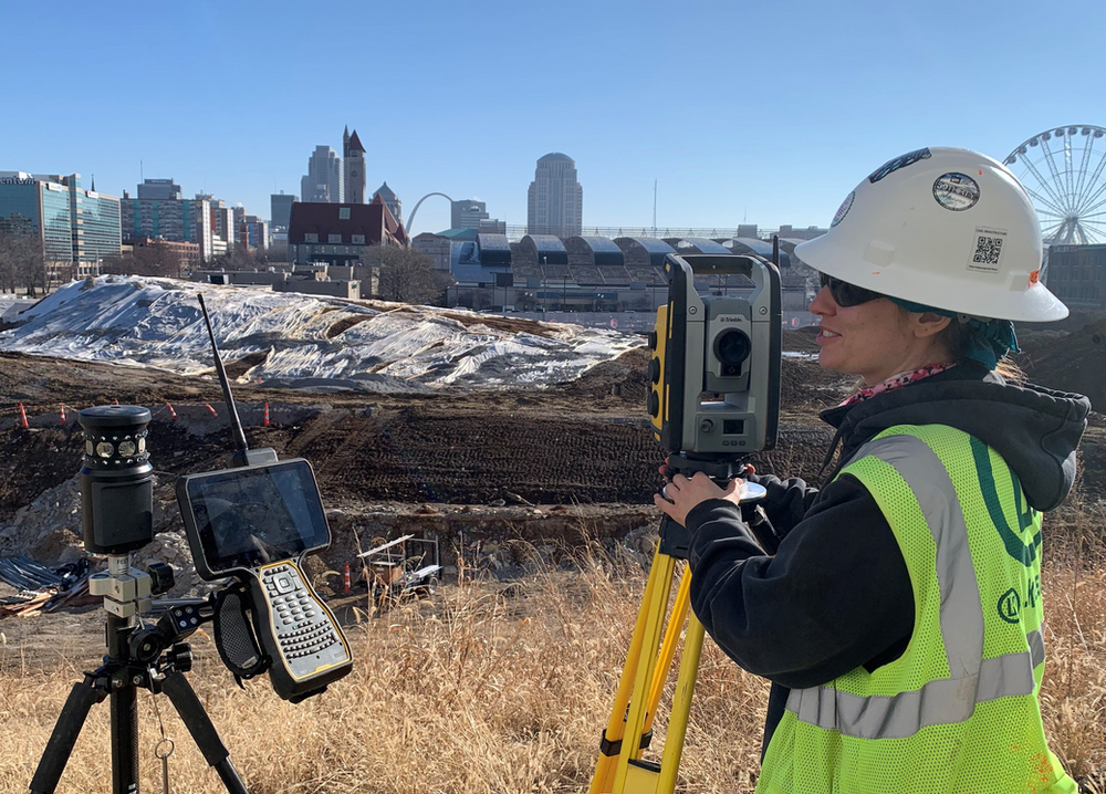 A woman in a hard hat and safety vest is using a total station.
