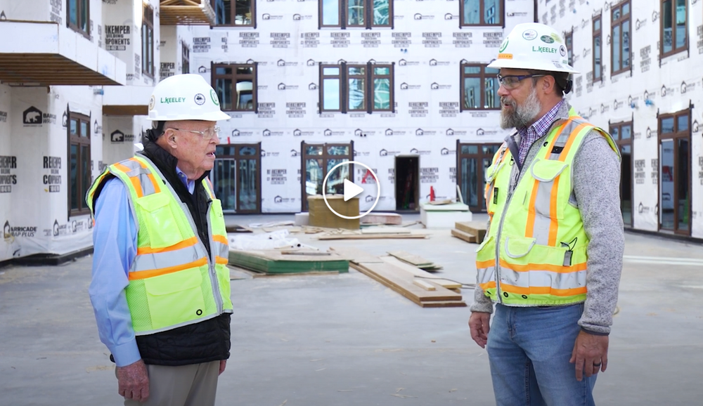 Two men wearing hard hats and safety vests are standing in front of a building under construction.