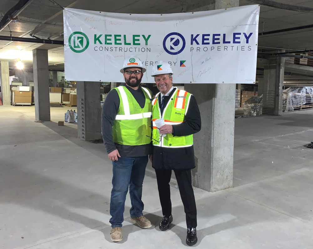 Two men are standing in front of a banner that says keeley construction