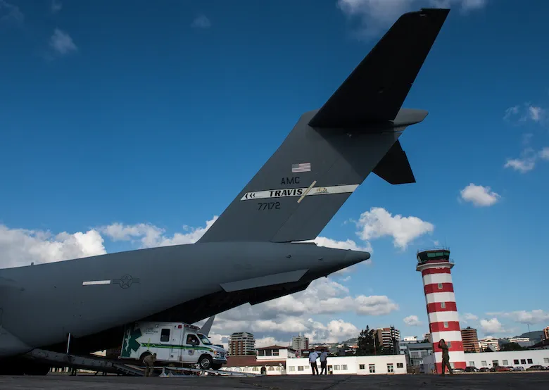 Military cargo plane on runway near a red-and-white control tower under a blue sky.
