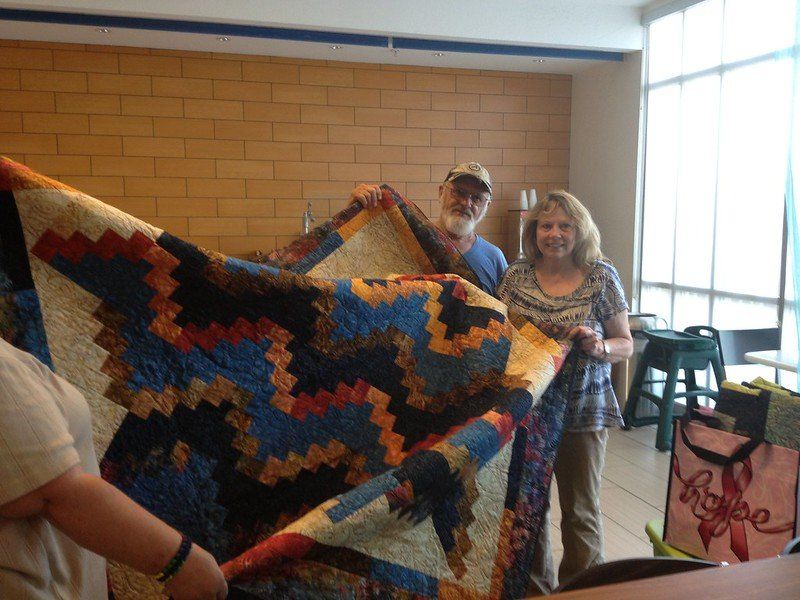 Two people hold up a colorful crocheted quilt in a bright room with windows and chairs.