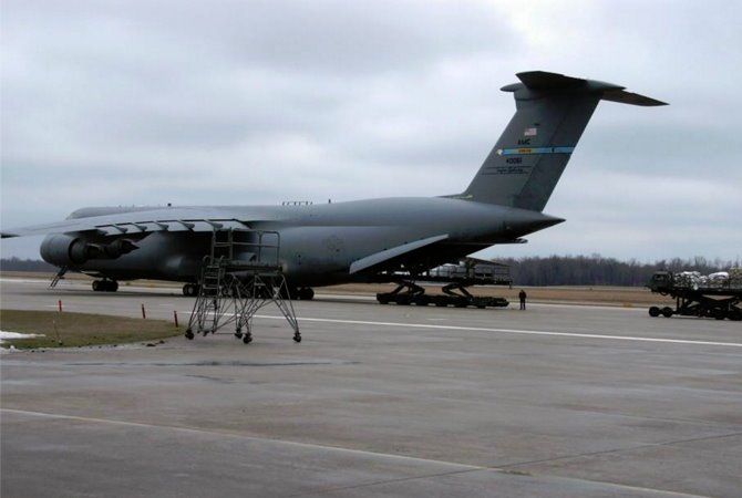 Gray military cargo plane on tarmac with landing gear and maintenance stands under overcast sky