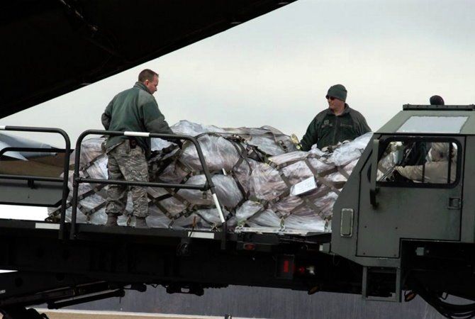 Workers unloading a truck bed piled with bundled packages wrapped in white plastic.