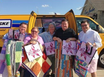 Five people holding quilt blankets and gift bags in front of a yellow van outdoors