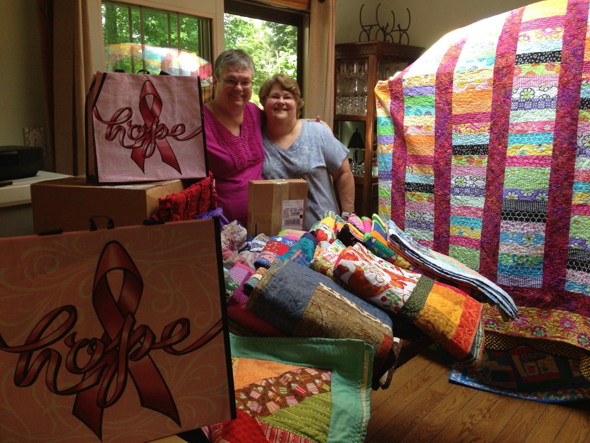 Two women standing beside colorful handmade quilts and gift bags in a bright room