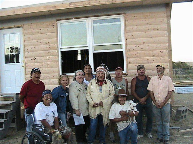 Group of people posing outside a wooden house with a large window and front door