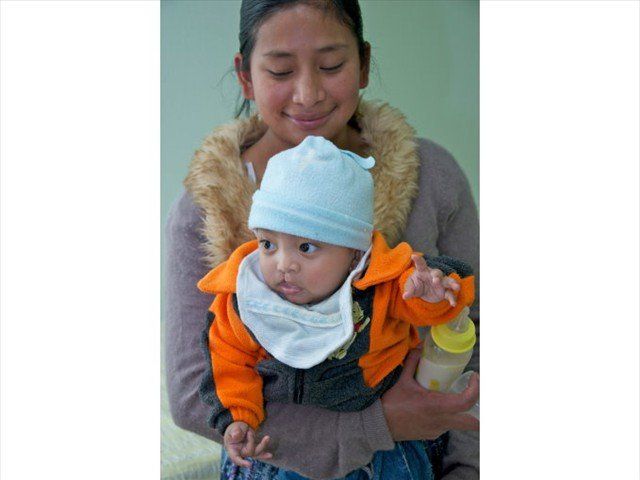 Woman holding a baby in an orange jacket and blue hat, both looking downward.
