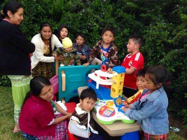 Children gathered around colorful toy tables outdoors, playing together by a hedge