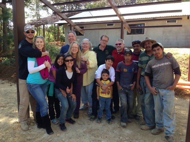 Group of people posing together outdoors under a wooden pavilion with trees and dry ground nearby.