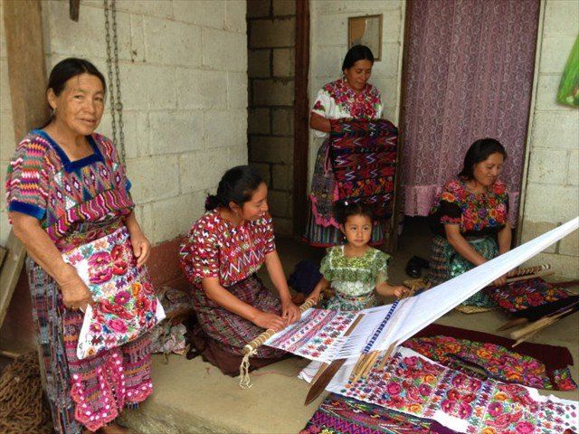 Four people in colorful embroidered clothing sit and stand in a doorway, weaving a long white textile.