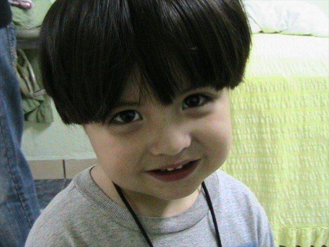 Close-up of a smiling child with dark hair, wearing a gray shirt and necklace, indoors by a bed.