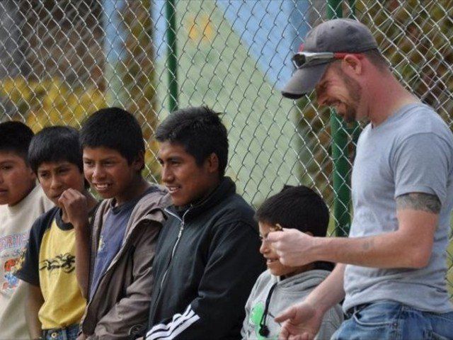 Man in gray cap hands food to children seated beside a chain-link fence outdoors