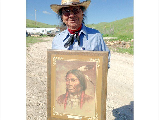 Man in a cowboy hat holding a framed portrait outdoors on a dirt road with green hills behind him