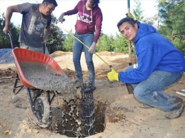Three people digging wet concrete with shovels beside a red wheelbarrow outdoors