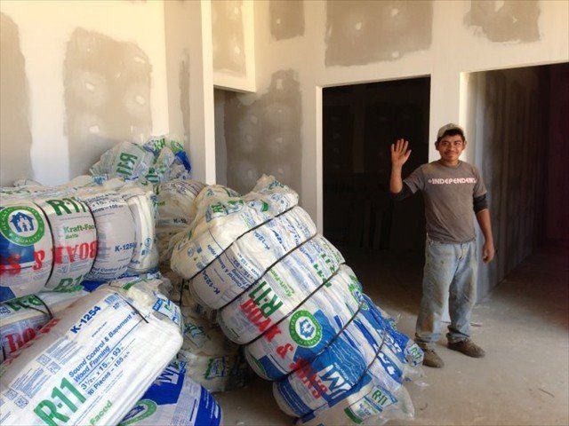 Man standing in an unfinished room beside stacks of wrapped insulation rolls, waving at the camera