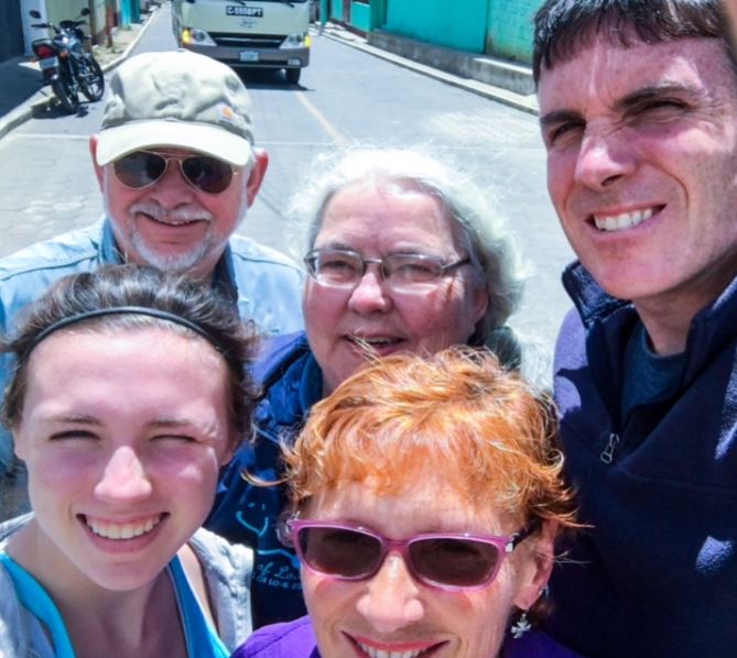 Five smiling people taking a selfie on a sunny street with cars and turquoise walls behind them