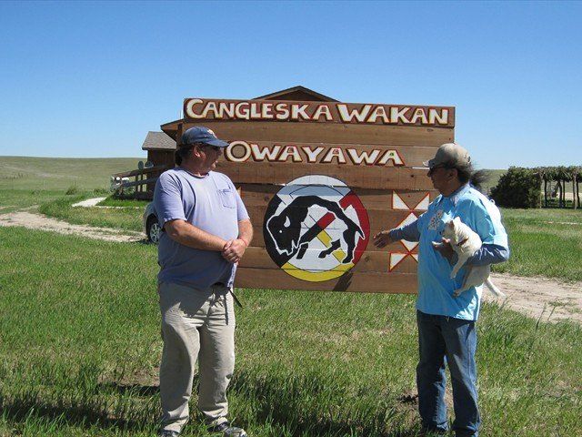 Two people standing by a Cangleska Wakan/Owayawa sign in a grassy field under a clear sky