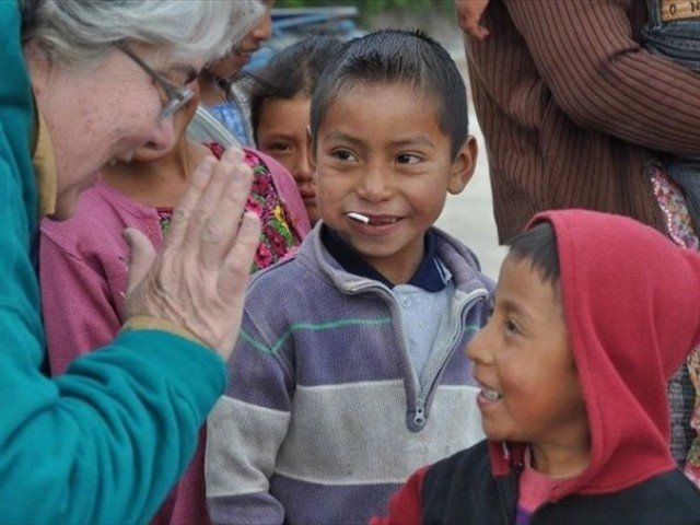 Woman smiles and greets two children outdoors, hands pressed together in a friendly gesture