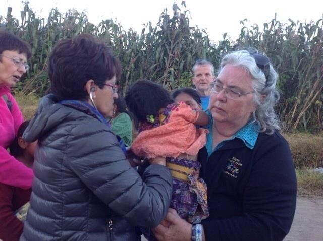 Several people outdoors sharing a baby wrapped in orange blanket on a rural path