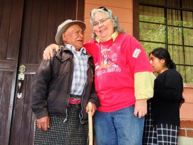 Two people smiling on a porch, one in a red shirt, with a child in the background