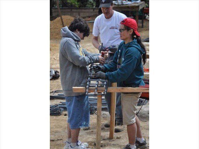 Three people handling tools at an outdoor workbench, with a man in a white shirt behind them.