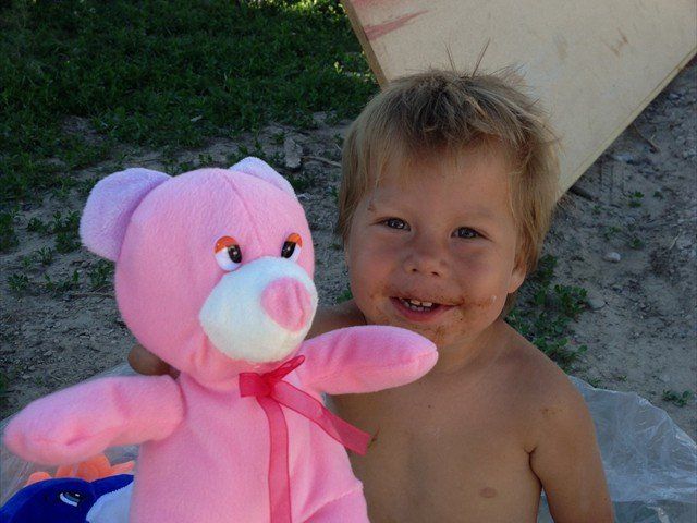 Smiling child holding a pink teddy bear outdoors on sandy ground